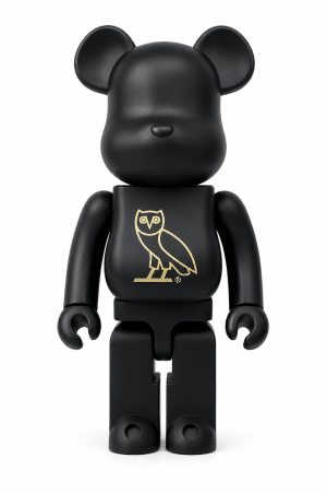 A matte black Bearbrick figure with a gold owl logo on its chest stands against a white background.