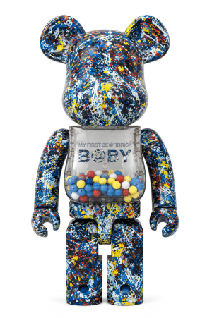 A Bearbrick figure covered in blue, yellow, red, and white paint splatters, with a clear torso showing "MY FIRST BE@RBRICK B@BY" and colorful beads.