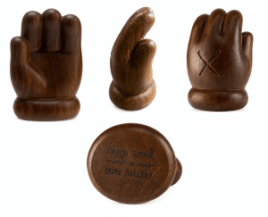 Four dark wooden sculptures of a cartoonish hand, one with an "X" carved into it, and one with "KAWS: GISWIL 12 JUNE - 28 AUGUST MORE GALLERY" engraved on its base.