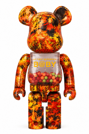 A Bearbrick figure with a clear torso filled with colorful candies and "B@BY" on its chest, covered in an orange and red maple leaf pattern.