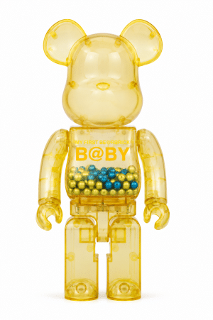 A transparent yellow Bearbrick toy stands, filled with gold and blue beads, with "MY FIRST BE@RBRICK B@BY" on its chest.